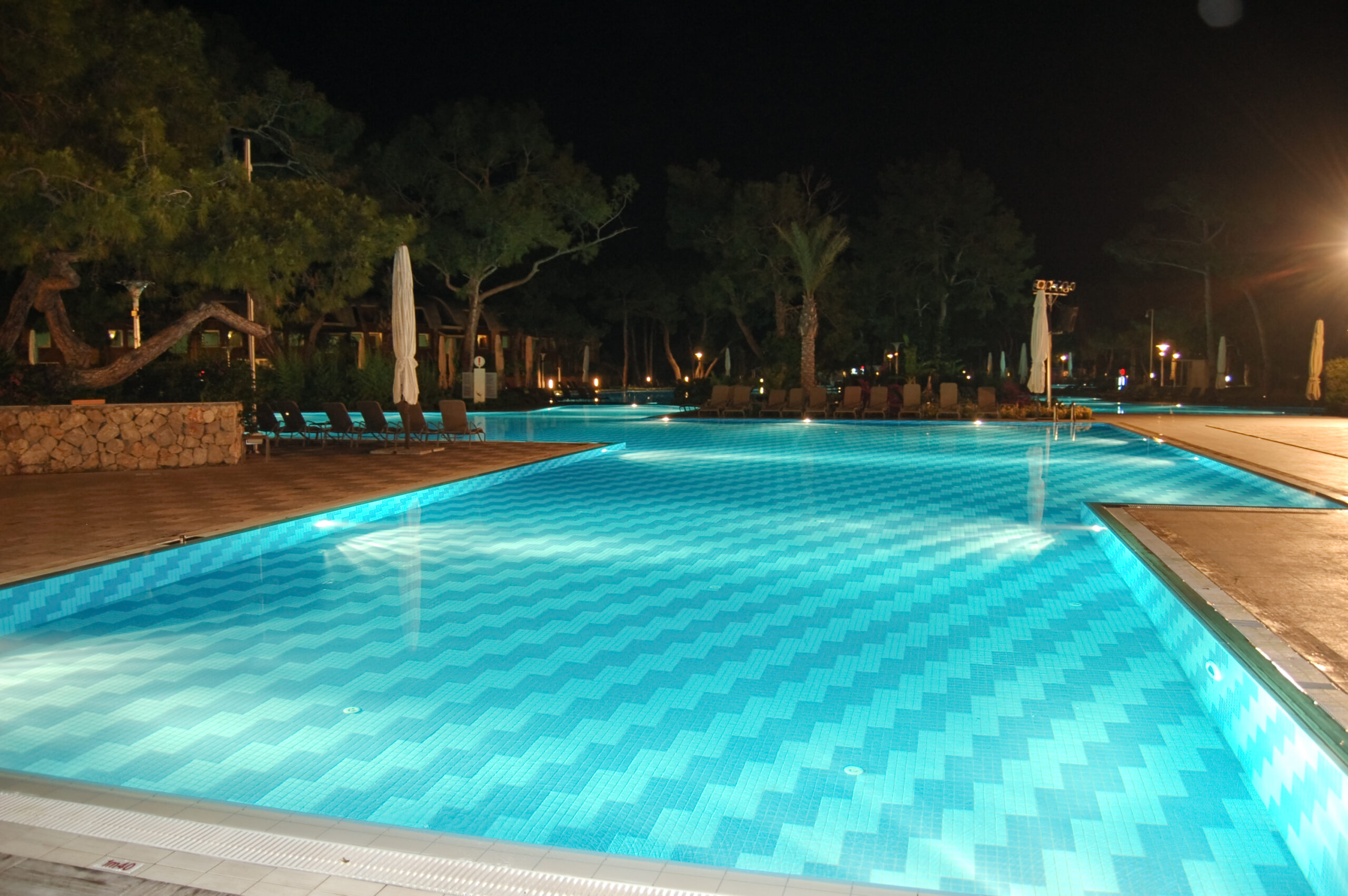 A nighttime view of a pool area surrounded by trees, with subtle lighting creating a tranquil and serene atmosphere