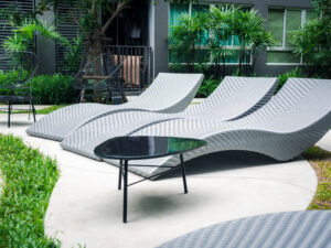 Modern grey curved rattan wicker sunbeds near outdoor swimming pool and green garden.