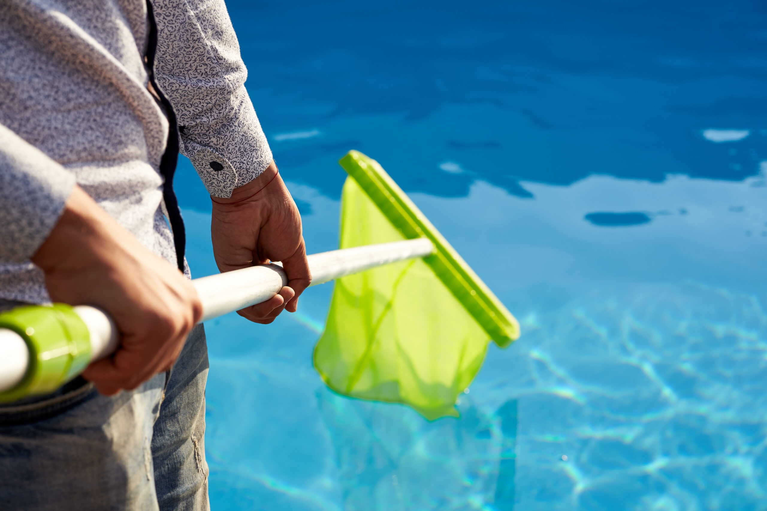 A man standing beside a swimming pool, holding a pool scoop net in his hand, ready to clean and remove debris from the pool water.